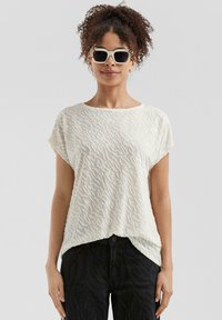 Blouse blanche texturée avec un motif ondulé, à manches courtes et coupe loose, associée à un jean noir. Modèle portant des lunettes de soleil.