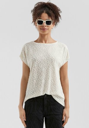 Blouse blanche texturée avec un motif ondulé, à manches courtes et coupe loose, associée à un jean noir. Modèle portant des lunettes de soleil.