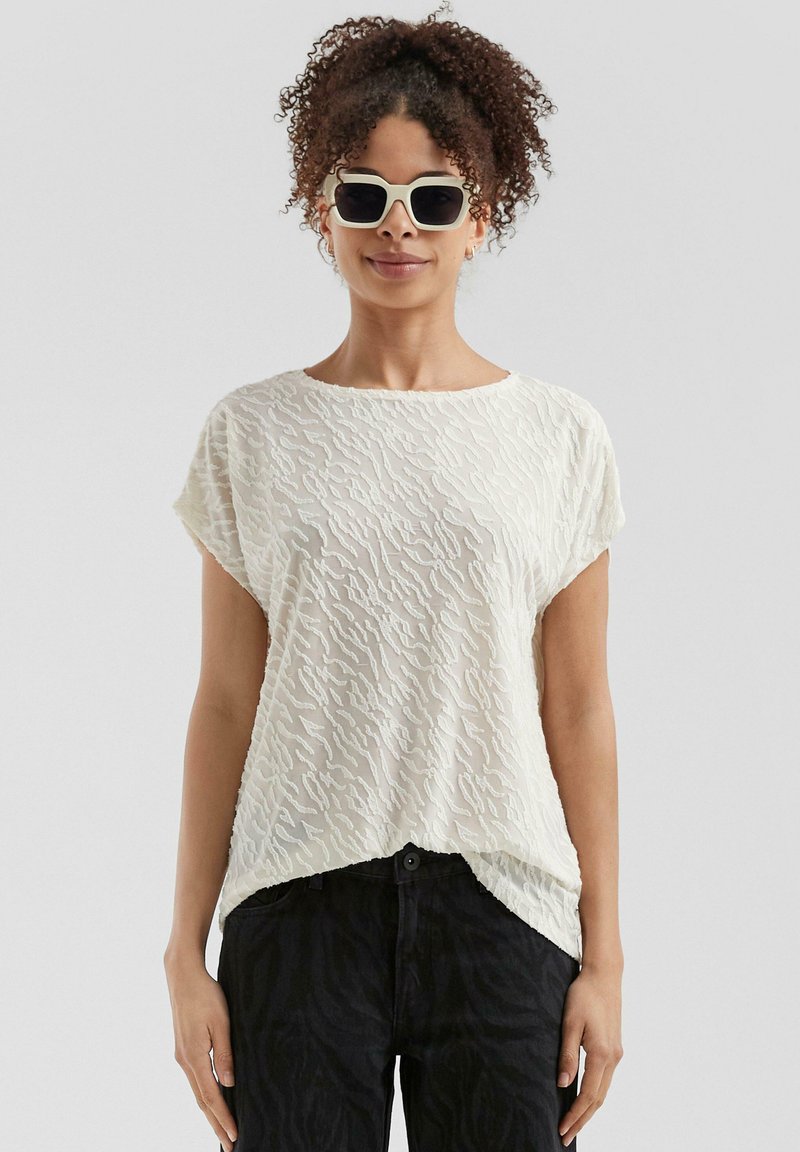 Blouse blanche texturée avec un motif ondulé, à manches courtes et coupe loose, associée à un jean noir. Modèle portant des lunettes de soleil.
