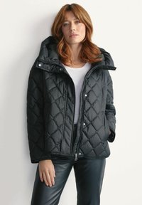 Czarna pikowana kurtka puchowa z kapturem, wyposażona w napy, zamek błyskawiczny oraz gładką teksturę. Nosi ją osoba w białej koszuli i ciemnych spodniach.