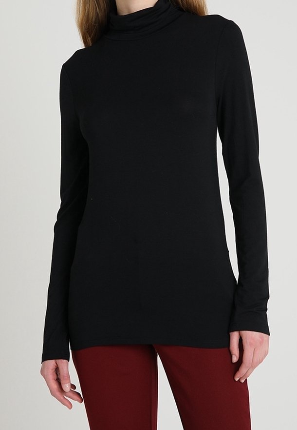 Femme portant un haut noir à col roulé à manches longues et un pantalon rouge foncé, debout devant un fond uni gris clair.
