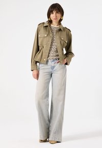 Olijfgroen cropped jack met zilveren knopen, twee voorzakken en een trekkoord in de taille, gecombineerd met lichtblauwe wijde jeans.