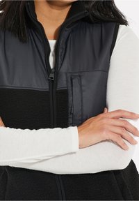 Chaleco de forro polar negro con un panel negro liso en la parte superior, que presenta un bolsillo con cremallera en el pecho y un cuello alto, llevado sobre una camisa blanca de manga larga.