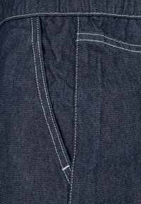 Tissu en denim bleu foncé avec des coutures blanches, présentant une poche latérale et une surface texturée. Le design met l'accent sur la durabilité et la praticité.