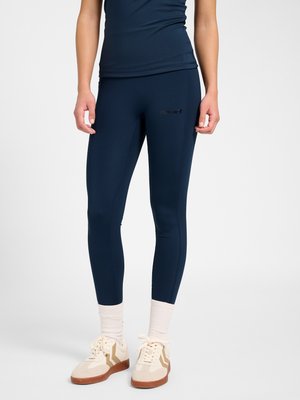 Person iført marineblå leggings, matchende top, hvide ankelsokker og beige sneakers med brune såler, stående mod en hvid baggrund.