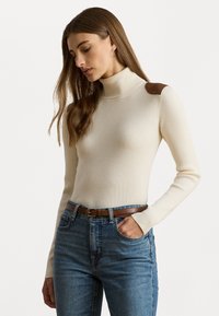 Ribbed cream turtleneck neulepaita, jossa on ruskeat olkaimet, yhdistettynä korkeavyötäröisiin sinisiin farkkuihin ja ruskeaan vyöhön.