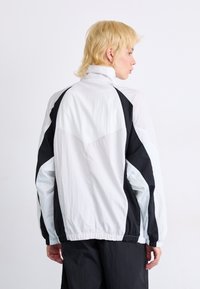 Veste coupe-vent blanche et noire avec un col montant, matériau texturé, poignets élastiques et panneaux contrastants sur les manches et le dos.