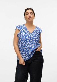 Blå blomstret bluse med korte volanger og V-hals. Stoffet har et struktureret mønster og en løs pasform, parret med mørke jeans.