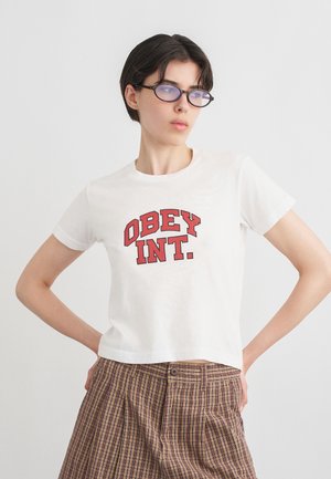Mladá osoba s krátkymi tmavými vlasmi nosí oválne okuliare, biele tričko s červenými písmenami "OBEY INT." a vysoký pás kockovaných nohavíc, ruky v bok.
