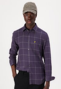 Purple checked shirt with a slim fit, button closure, and long sleeves. Features a small yellow logo embroidered on the chest. Worn with a gray cap.
