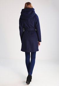 Marinblå ullkappa med stor huva, midjebälte och långa ärmar, kombinerad med åtsittande mörkblå byxor och svarta ankelstövlar.