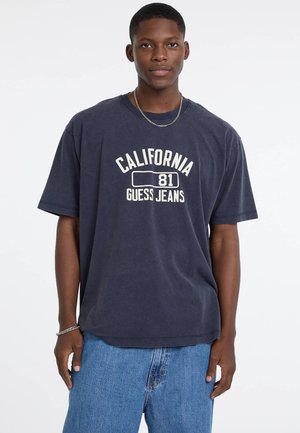 Granatowa, oversize'owa koszulka z krótkim rękawem z napisem "CALIFORNIA 81 GUESS JEANS" w kolorze białym. Okrągły dekolt, krótki rękaw, miękka bawełniana tkanina. Widać dżinsowe szorty.
