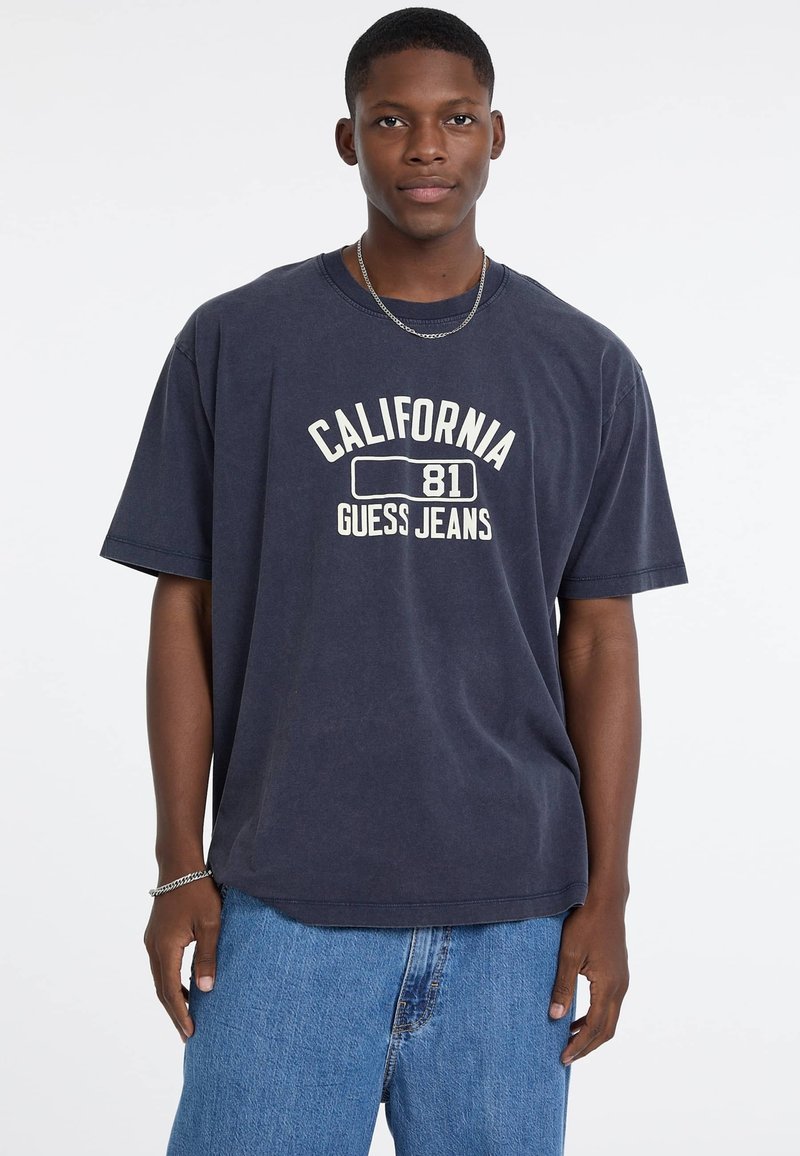 Granatowa, oversize'owa koszulka z krótkim rękawem z napisem "CALIFORNIA 81 GUESS JEANS" w kolorze białym. Okrągły dekolt, krótki rękaw, miękka bawełniana tkanina. Widać dżinsowe szorty.