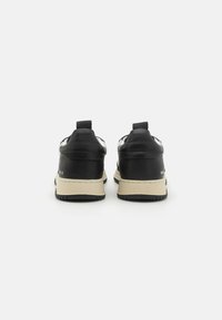 Baskets en cuir noir avec semelles en caoutchouc beige, présentant une finition texturée et une languette à l'arrière pour un enfilage facile. Pas de logos visibles.