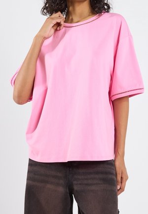 Kobieta ubrana w luźną, różową, krótką bluzkę z przeszyciami na kołnierzu i rękawach, zestawioną z ciemnymi, przetartymi dżinsami, na jednolitym tle.