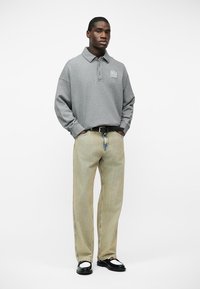 Νεαρός άνδρας με γκρι μακρυμάνικο πόλο πουκάμισο, μπεζ τζιν με φαρδιά πόδια και μαύρα-άσπρα παπούτσια, στέκεται με τα χέρια στις τσέπες.