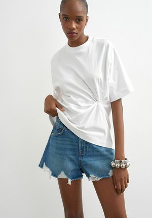 Vrouw draagt een wit t-shirt met een gedraaide voorkant, versleten spijkershorts en een dikke zilveren armband aan haar rechterpols.