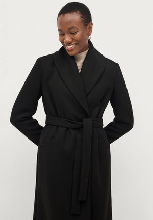 Smiling woman with short hair wears black belted coat over beige ribbed turtleneck against plain light background.
