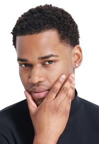 Gros plan sur une personne avec les cheveux courts et bouclés, portant un haut noir. Ils ont un teint lisse et une boucle d'oreille en diamant dans l'oreille gauche.