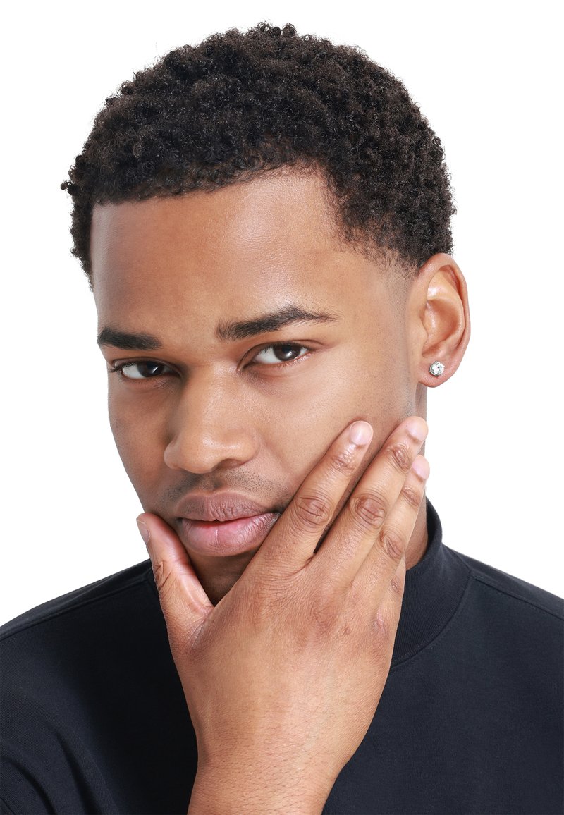 Gros plan sur une personne avec les cheveux courts et bouclés, portant un haut noir. Ils ont un teint lisse et une boucle d'oreille en diamant dans l'oreille gauche.