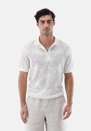 Junger Mann trägt ein weißes, kurzärmeliges Strick-Poloshirt mit Palmenmuster und hellbeige Hose, steht vor einem schlichten Hintergrund.