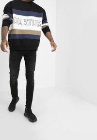 Svart och blå randig sweatshirt med stor text, i kombination med slitna svarta jeans och svarta sneakers. Slät textur, lös passform.