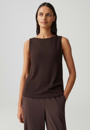 Frau mit langen dunklen Haaren, die ein ärmelloses, dunkelbraunes Oberteil und passende lockere Hosen trägt, steht mit einer Hand in der Tasche.