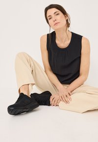 Svart ärmlös topp med plisserad halsringning, kombinerad med beige vida byxor och svarta sneakers, sittande mot en neutral bakgrund.