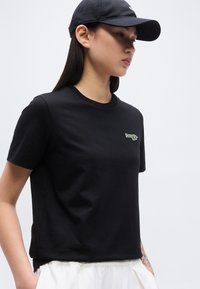 Νεαρή γυναίκα που φοράει μαύρο καπέλο και μαύρο κοντομάνικο μπλουζάκι με πράσινο και λευκό λογότυπο, συνδυασμένο με λευκό παντελόνι.