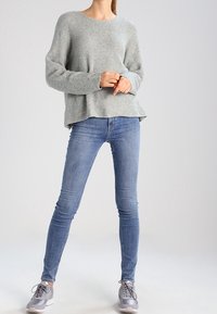 Suéter de punto gris claro con mangas largas, escote redondo y un corte holgado, combinado con jeans ajustados azules y zapatillas grises.