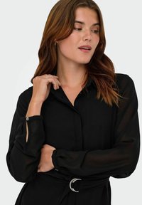 Czarna sukienka koszulowa z długim rękawem, kołnierzykiem, przezroczystymi rękawami, zapinanymi mankietami oraz paskiem z okrągłą klamrą w odcieniu srebrnym.
