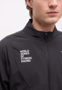 Νεαρό άτομο που φοράει μαύρο μπουφάν με φερμουάρ, με την ένδειξη «World Series of Fitness Racing» τυπωμένη σε λευκά γράμματα στο στήθος.