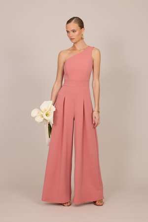 Γ γυναίκα με αμάνικη, μονό ωμο ολόσωμη φόρμα σε σκουρό ροζ χρώμα κρατά λευκά κάλλα λίλιουμ, στέκεται μπροστά σε απλό μπεζ φόντο.