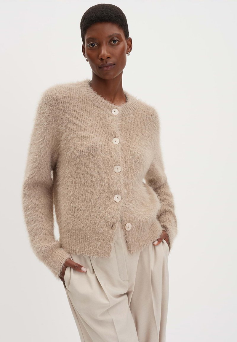 Gilet beige en peluche avec un col rond, doté de cinq boutons crème et de poignets côtelés. Porté sur un pantalon de couleur claire.