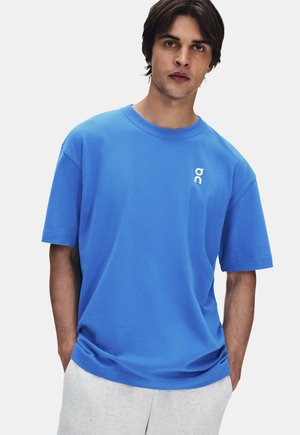 Mann trägt ein leuchtend blaues kurzärmeliges T-Shirt mit kleinem weißem Logo auf der linken Brust und hellgraue Hose.