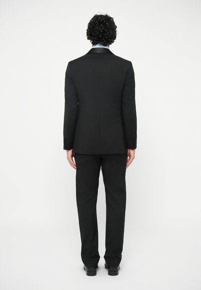 Osoba s kudrnatými vlasy stojí zády, oblečená v černém obleku, světle modré košili a černých botách na jednobarevném bílém pozadí.