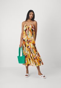 Maxi dress with a colorful abstract pattern in orange, pink, and brown. Features spaghetti straps and a ruffled hem. Holding a green bag.