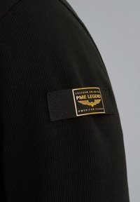 Czarna prążkowana tkanina z złotą i czarną metką, na której znajdują się napisy "PME Legend", "Freedom Unlimited" oraz akcenty "American Classic".