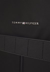 Tessuto nero con una finitura testurizzata, ornato da un logo metallico "TOMMY HILFIGER" e un dettaglio a tracolla orizzontale nella parte inferiore.