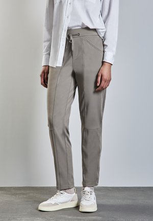 Pantalons gris clair fuselés avec une texture lisse, présentant des coutures inclinées, une taille à cordon de serrage, et accompagnés de baskets blanches.