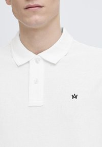 Biely polo tričko s textúrovanou tkaninou, má klasický golier, dvojité zapínanie na gombíky a malé čierne logo na ľavej strane hrudníka.