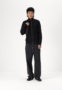 Maglione nero a coste con zip e colletto alto, abbinato a jeans neri dal taglio largo e scarpe nere. Porta una piccola borsa nera.