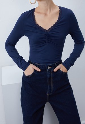 Femme en haut bleu marine à manches longues et col en V avec bordure en dentelle, rentré dans un jean en denim foncé taille haute, mains dans les poches, portant des boucles d'oreilles anneaux dorés.