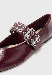 Burgundowe lakierowane buty ze zaokrąglonym noskiem, z szerokim paskiem ozdobionym srebrnymi oczkami i metalowym zapięciem.