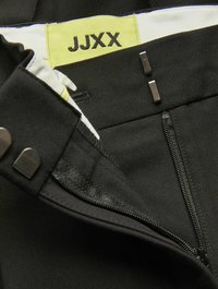 Črne hlače z zadrgo in kovinskim zapiranjem, z rumenim oznako "JJXX" prišito. Gladek material z rahlim sijajem.