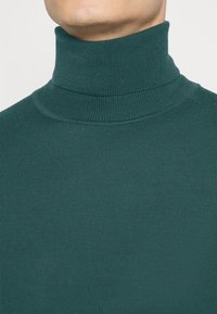 Grüner gerippter Rollkragenpullover mit hohem Kragen, der ein strukturiertes Gewebe aufweist. Das Material ist weich und figurbetont.