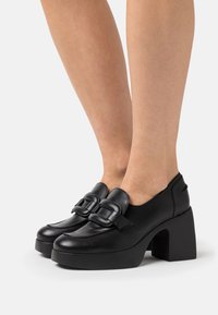 Zapatos de plataforma de cuero negro con punta redonda, que presentan detalles decorativos con hebillas y un grueso tacón cuadrado. Textura suave.