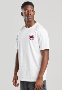 Valkoinen puuvillapaita, jossa on pieni punainen ja musta grafiikkasuunnittelu säteilevin viivoin vasemmassa rinnassa, pyöreä kaula-aukko ja lyhyet hihat.