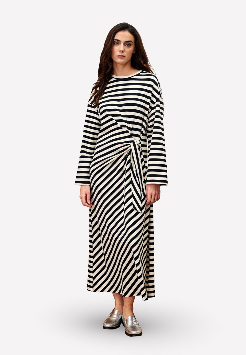 Frau in einem langen schwarz-weißen gestreiften Kleid mit einem geknoteten Detail in der Taille und metallisch silbernen Slippers, die vor einem weißen Hintergrund steht.
