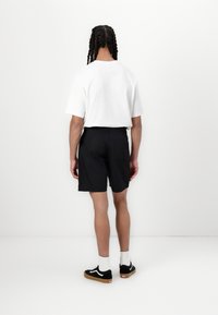 Chemise blanche à manches courtes, shorts noirs et baskets noires avec semelles en gomme. Le modèle se tient de dos face à un fond uni.